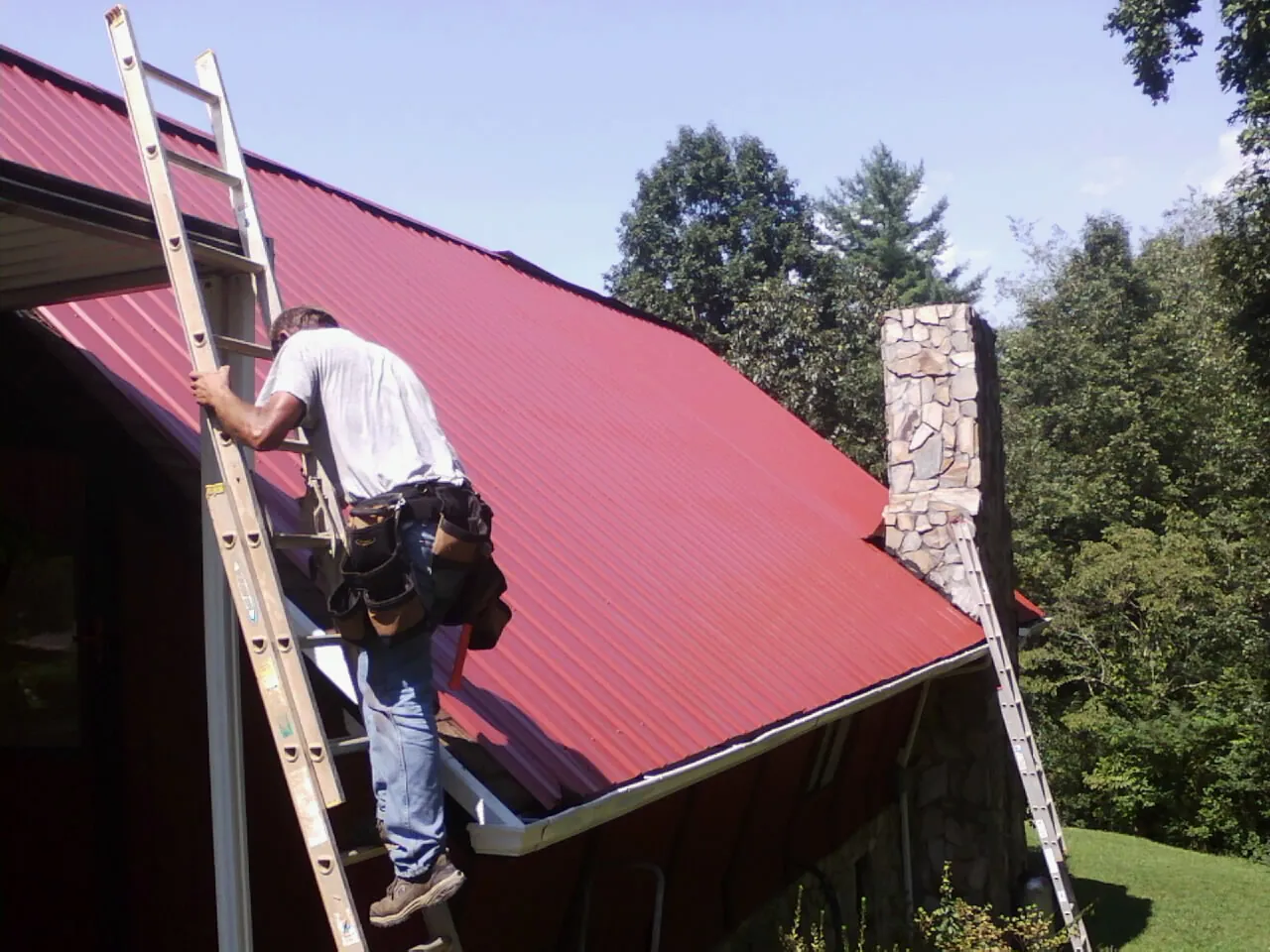 Roofer on a ladder beside a red metal roof and stone chimney — Soffit & Fascia Repair by Roofing Brothers in Springboro